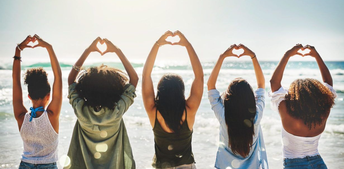 Five women on a beach making heart shapes with their hands